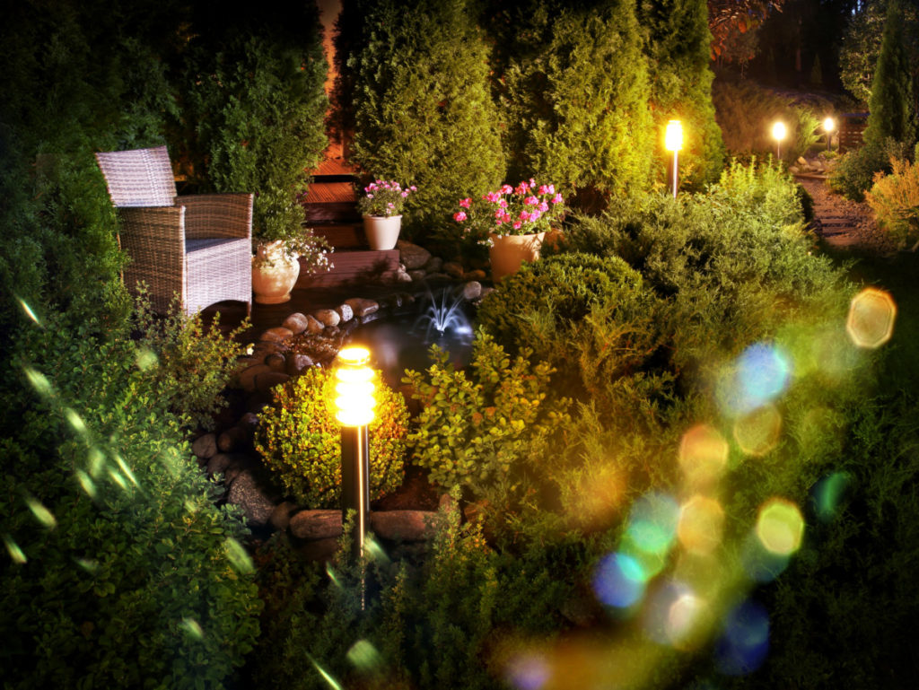 A backyard patio with colorful lights and a small decorative pond in the center. It's evening and everything is softly lit. There are small trees in the background and a chair next to the pond.