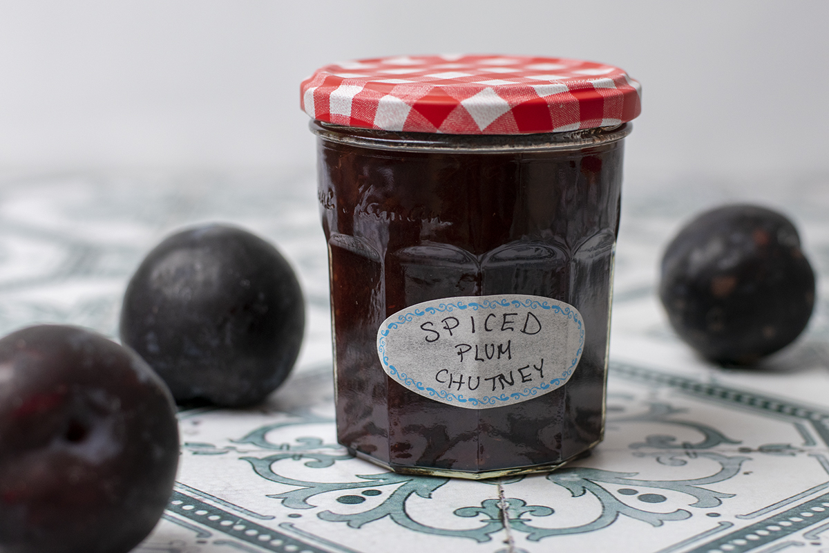 A jar of spiced plum chutney on a tiled surface with several plums